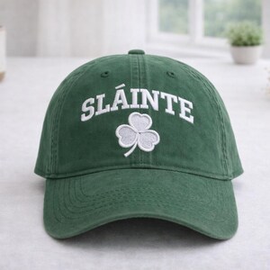 May include: A green baseball cap featuring the word "SLÁINTE" in white lettering and a white shamrock. The hat has a curved peak and is made of a soft fabric. This casual cap is a stylish accessory.