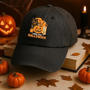 May include: A dark gray baseball cap with a Halloween-themed graphic. The graphic includes a black cat, a carved pumpkin, and the text "LONG LIVE HALLOWEEN" in orange. The cap is surrounded by pumpkins and autumn leaves.