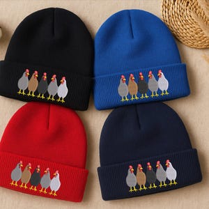Könnte beinhalten: Vier Strickmützen in Schwarz, Blau, Rot und Marine. Jede Mütze hat eine gestickte Reihe von Hühnern in verschiedenen Farben. Die Mützen werden auf einer neutralen Oberfläche mit dekorativen Elementen präsentiert.
