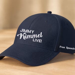 Può includere: Un cappellino da baseball blu navy con la scritta bianca "JIMMY Kimmel LIVE!". La frase "Free Speech" è stampata sul lato. Appoggiato su una superficie di legno.