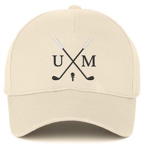 May include: A cream-coloured baseball cap with a curved peak. The cap displays a design of crossed golf clubs with the letters "U" and "M" on either side, and a golf ball and tee in the centre. The cap is made of a soft fabric.