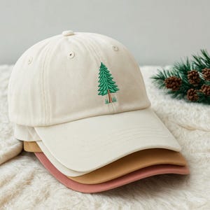 Può includere: Una pila di quattro cappellini da baseball in colori neutri. Il cappellino superiore è bianco sporco con un disegno di albero ricamato verde. Gli altri cappellini sono beige e rosa. I cappellini sono realizzati in tessuto morbido e hanno una visiera curva.