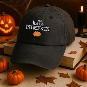 Könnte beinhalten: Dunkelgraue Baseballkappe mit dem Schriftzug "hello PUMPKIN" in Weiß und einer orangefarbenen Kürbis-Illustration. Das Bild zeigt Kürbisse, Herbstblätter und eine brennende Kerze, die eine herbstliche Atmosphäre erzeugt.