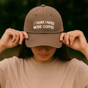 Peut inclure: Casquette marron avec le texte "I THINK I NEED MORE COFFEE" en blanc. La casquette est tenue par une personne. La personne porte un t-shirt beige.