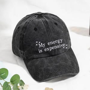 Könnte beinhalten: Verwaschene schwarze Baseballkappe mit dem weißen Schriftzug "My energy is expensive". Die Kappe hat einen gebogenen Schirm und eine runde Krone. Der Text ist von kleinen sternförmigen Designs umgeben.