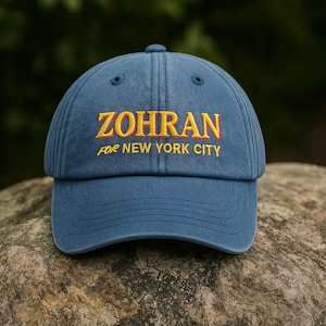Könnte beinhalten: Eine marineblaue Baseballkappe mit dem gestickten Schriftzug "ZOHRAN" in Gelb und Rot, darunter "FOR NEW YORK CITY". Die Kappe liegt auf einem Stein.