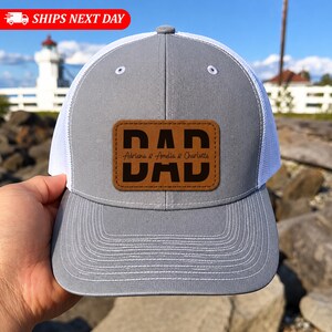 Könnte beinhalten: Graue Trucker-Mütze mit weißem Mesh-Rücken und einem braunen Lederaufnäher. Der Aufnäher zeigt das Wort "DAD" in fetten schwarzen Buchstaben, darunter die Namen "Adriana & Amelia & Charlotte". Die Mütze hat einen gebogenen Schirm.