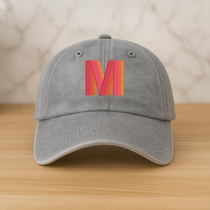 Könnte beinhalten: Graue Baseballkappe mit einem farbenfrohen, gestickten "M" in Rot, Orange und Gelb. Die Kappe ist aus einem weichen Stoff gefertigt und hat einen gebogenen Schirm. Der Hut steht auf einer Holzoberfläche.