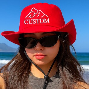 Könnte beinhalten: Ein roter Sonnenhut mit weißem Bergmotiv und dem Wort "CUSTOM" auf der Vorderseite. Der Hut hat eine breite Krempe und einen schwarzen Kinnriemen. Die Person trägt eine schwarze Sonnenbrille.