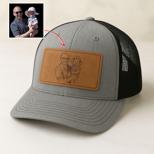 Könnte beinhalten: Graue Trucker-Kappe mit schwarzem Mesh-Rücken und einem braunen Lederpatch. Der Patch zeigt eine Schwarz-Weiß-Skizze einer Person, die ein Kind hält. Die Kappe hat einen gebogenen Schirm und einen Druckknopfverschluss.