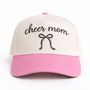 Könnte beinhalten: Baseballcap mit cremefarbener Krone und pinkfarbenem Schirm. Die Worte "cheer mom" sind in schwarzer Schrift über einem schwarzen Schleifendesign gestickt. Die Kappe hat einen rosa Knopf oben.