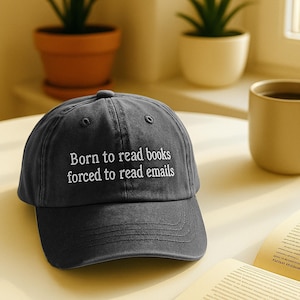 May include: A dark grey baseball cap with white embroidered text reading "Born to read books forced to read emails". The cap is on a yellow surface, with a coffee mug and a book visible in the background.
