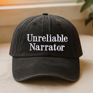 Könnte beinhalten: Eine schwarze Baseballkappe mit dem weißen Schriftzug "Unreliable Narrator". Die Kappe hat einen gebogenen Schirm und einen Knopf oben. Das Material ist weich und leicht verwaschen.