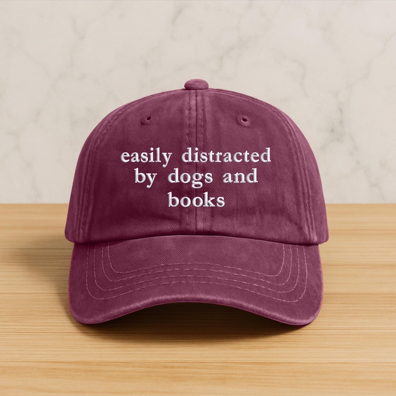 Może przedstawiać: Bordowa czapka z daszkiem z białym napisem "easily distracted by dogs and books". Czapka ma zakrzywiony daszek i zapięcie na pasek z tkaniny.