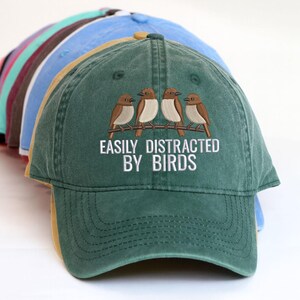 Könnte beinhalten: Eine Sammlung von Baseballkappen in verschiedenen Farben, wobei der Fokus auf einer grünen Kappe liegt. Die grüne Kappe hat ein gesticktes Design von drei Vögeln auf einem Ast mit dem weißen Text "EASILY DISTRACTED BY BIRDS".