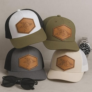 May include: Four baseball caps in different colours: white, black, olive green, and grey. Each cap has a leather patch with a hand-drawn design and the words "Daddy" or "Papa". Sunglasses and a wristwatch are also visible.