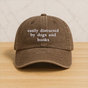 Może przedstawiać: Brązowa czapka bejsbolowa z białym haftem "easily distracted by dogs and books". Czapka ma zakrzywiony daszek i zaokrągloną koronę. Kapelusz jest wykonany z miękkiego, teksturowanego materiału.