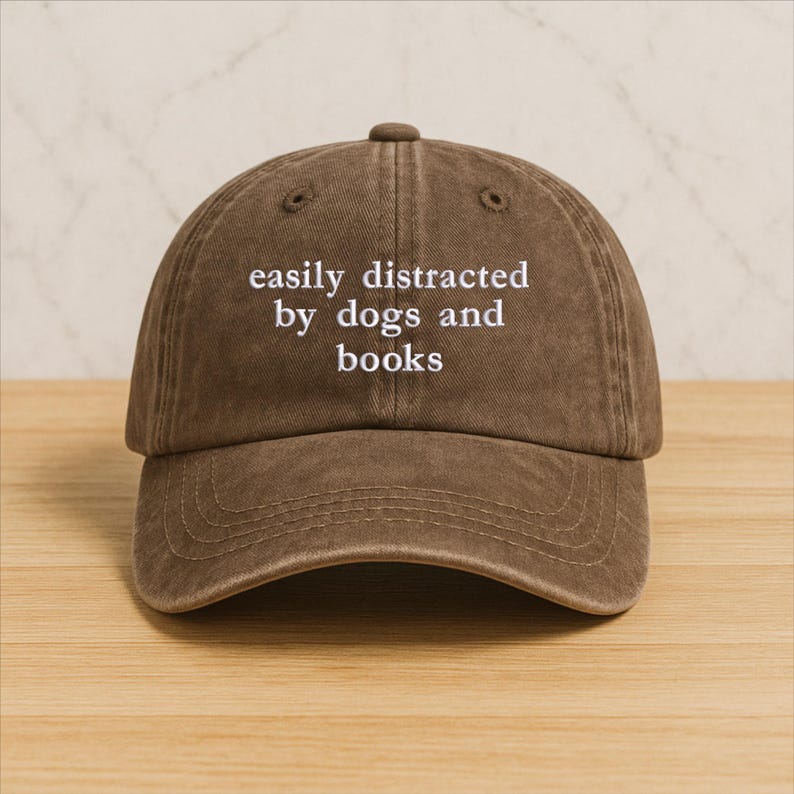 Może przedstawiać: Brązowa czapka bejsbolowa z białym haftem "easily distracted by dogs and books". Czapka ma zakrzywiony daszek i zaokrągloną koronę. Kapelusz jest wykonany z miękkiego, teksturowanego materiału.