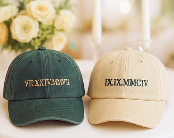 May include: Two baseball caps, one dark green and one tan, sit side-by-side. The dark green cap has gold Roman numerals embroidered on the front. The tan cap has black Roman numerals embroidered on the front. The caps are on a white surface.