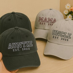 May include: Four baseball caps in shades of gray and black are displayed. One black cap reads "AUNTIE EST. 2025". A light gray cap says "UNCLE EST. 2025". Another light gray cap says "MAMA EST. 202" and a black cap is in the background.