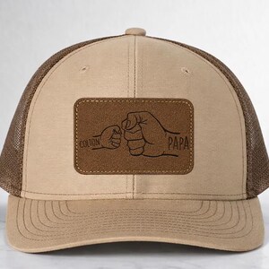 Könnte beinhalten: Beige und braune Trucker-Cap mit Mesh-Rückseite. Auf der Vorderseite befindet sich ein braunes Lederpatch mit den Wörtern "Colton" und "Papa" über einer Faust-Illustration.