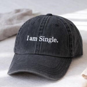 Op de afbeelding: Een donkergrijze baseballcap met de geborduurde tekst "I am Single." De pet heeft een gebogen klep en een stoffen bandsluiting. Een casual accessoire.