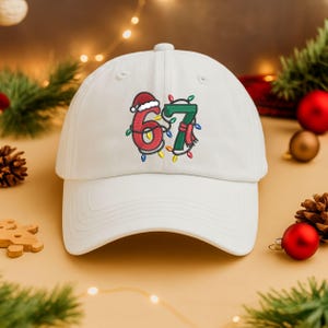 Könnte beinhalten: Weiße Baseballkappe mit einem festlichen Stickdesign. Das Design zeigt die Zahlen "67", die mit Weihnachtslichtern, einem Weihnachtsmannhut und einem Schal verziert sind. Ein lustiges und saisonales Accessoire.