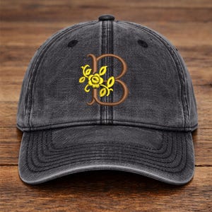 Könnte beinhalten: Dunkelgraue Baseballkappe mit gebogenem Schirm. Die Kappe hat einen gestickten braunen Buchstaben "B" mit einem gelben Blumendesign. Die Kappe hat einen Used-Look.