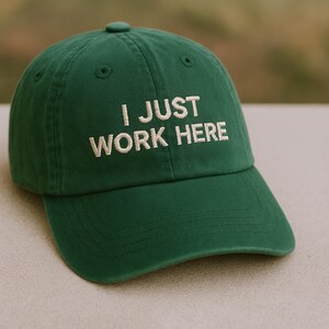 Könnte beinhalten: Eine grüne Baseballkappe mit dem gestickten Text "I JUST WORK HERE" in Weiß. Die Kappe hat einen gebogenen Schirm und einen Knopf oben. Der Hut ist aus einem weichen Stoff gefertigt.