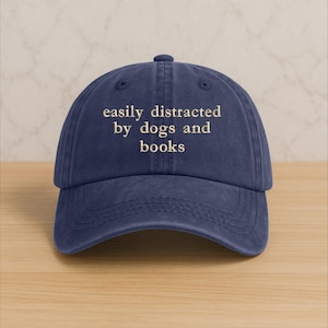 Może przedstawiać: Granatowa czapka z daszkiem z haftowanym kremowym napisem "easily distracted by dogs and books". Czapka ma zakrzywiony daszek i zapięcie na pasek z tkaniny.