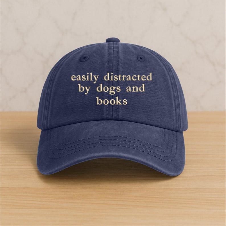 Może przedstawiać: Granatowa czapka z daszkiem z haftowanym kremowym napisem "easily distracted by dogs and books". Czapka ma zakrzywiony daszek i zapięcie na pasek z tkaniny.