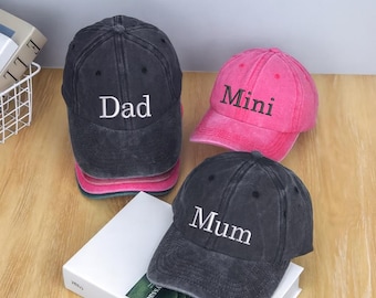 May include: Three baseball caps in the image. One is black with "Dad" in white, another is pink with "Mini" in white, and the third is black with "Mum" in white. The caps are on a wooden surface.