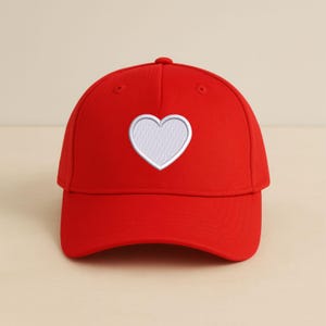 Könnte beinhalten: Rote Baseballkappe mit einem weißen Herz, das auf der Vorderseite gestickt ist. Die Kappe hat einen gebogenen Schirm und eine abgerundete Krone. Das Herz ist weiß umrandet und mit weißen Stichen gefüllt. Die Kappe ist aus glattem Stoff.