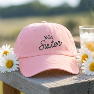 Könnte beinhalten: Hellrosa Baseballkappe mit dem gestickten Text "Big Sister" in dunkelgrauer Schrift. Die Kappe liegt auf einer Holzoberfläche neben weißen Gänseblümchen mit gelben Zentren.