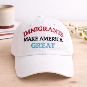 Könnte beinhalten: Weiße Baseballkappe mit dem gestickten Text "IMMIGRANTS MAKE AMERICA GREAT" in Rot, Schwarz und Blau. Die Kappe liegt auf einer hellen Holzoberfläche, im Hintergrund eine Tasse und Bücher.
