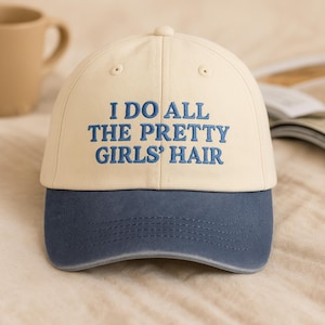 Peut inclure: Casquette de baseball avec une couronne crème et une visière bleu marine. Le texte brodé en bleu "I DO ALL THE PRETTY GIRLS' HAIR" est visible. Un accessoire décontracté.