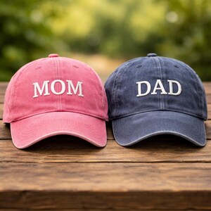 Könnte beinhalten: Zwei Baseballkappen, eine rosa und eine marineblaue, liegen nebeneinander auf einer Holzoberfläche. Die rosa Kappe hat das Wort "MOM" in Weiß gestickt, während die marineblaue Kappe "DAD" in Weiß gestickt hat.