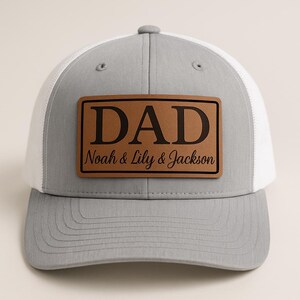 Könnte beinhalten: Grau-weiße Trucker-Cap mit einem braunen Lederaufnäher. Der Aufnäher zeigt das Wort "DAD" in fetten, schwarzen Buchstaben, darunter "Noah & Lily & Jackson" in Schreibschrift. Die Cap hat einen gebogenen Schirm und eine Mesh-Rückseite.
