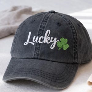 Könnte beinhalten: Dunkelgraue Baseballkappe mit dem Wort "Lucky" in weißer Schrift, begleitet von einem grünen Kleeblatt. Die Kappe hat einen gebogenen Schirm und einen Used-Look, der einen Vintage-Stil suggeriert.