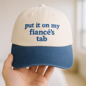 Może przedstawiać: Czapka bejsbolowa z kremową koroną i granatowym daszkiem. Czapka ma wyhaftowany napis "put it on my fiancé's tab" w granatowym kolorze. Casualny dodatek.