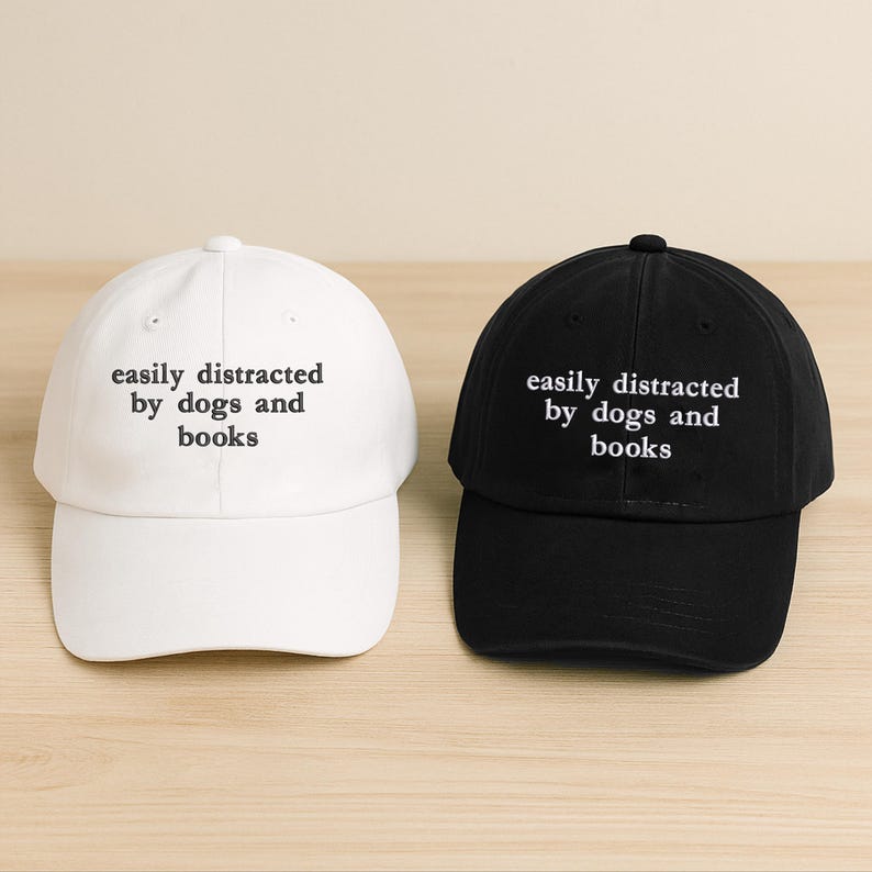 Może przedstawiać: Dwie czapki bejsbolowe, jedna biała i jedna czarna, są pokazane. Obie czapki mają wyhaftowany biały napis "easily distracted by dogs and books". Czapki leżą na jasnej powierzchni imitującej drewno.