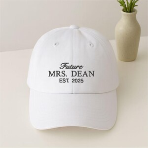 May include: A white baseball cap featuring the text "Future MRS. DEAN EST. 2025" in black script. The cap has a curved peak and a button on top. A decorative vase with greenery is in the background.