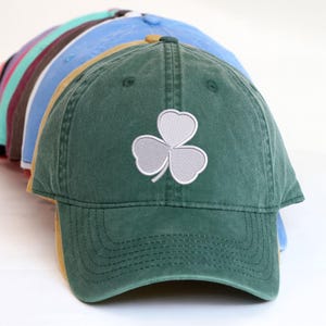 Könnte beinhalten: Eine Auswahl an Baseballkappen in verschiedenen Farben, darunter Grün, Blau und Rosa. Die vorderste Kappe ist grün und hat ein weiß-graues Kleeblatt-Emblem. Die Kappen sind in einer Reihe angeordnet und zeigen ihre unterschiedlichen Farbtöne und Designs.
