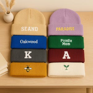 Könnte beinhalten: Eine Auswahl an gerippten Strickmützen in verschiedenen Farben, darunter Gelb, Blau, Grau und Burgunderrot. Jede Mütze hat gestickten Text oder eine kleine Grafik, wie "SEAND", "Oakwood", "K" und eine Biene. Die Mützen sind auf einer Holzoberfläche gestapelt.