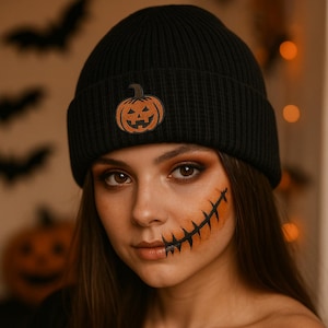 Könnte beinhalten: Eine schwarze Strickmütze mit einem braunen Kürbis-Patch. Die Mütze wird von einer Person mit Halloween-Make-up getragen, einschließlich eines aufgenähten Munddesigns auf der Wange. Der Hintergrund enthält Halloween-Dekorationen.