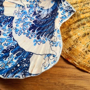 May include: Close-up of two decorative seashells. One shell features a blue and white wave pattern, while the other is gold with glitter accents. The shells are on a wooden surface, showcasing their unique textures and designs.