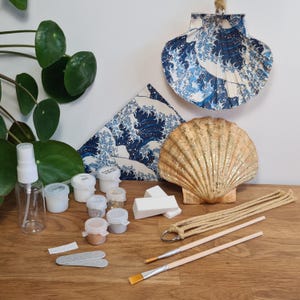 May include: A selection of craft materials and decorative objects on a wooden surface. Features a large, gold seashell, small paint pots, brushes, and a hanging seashell ornament with a blue and white wave design. Also includes a spray bottle, rope, and nail files.
