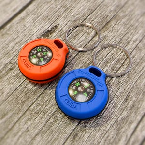 May include: Two compass keychains, one orange and one blue, with silver key rings. The compasses have a black dial with white and green markings. The brand name "CEIBA" is printed on the compass face.