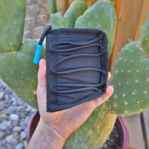 Puede incluir: Una cartera negra con detalles de cordón elástico gris y una lengüeta azul. La cartera se sostiene frente a un cactus, destacando su diseño compacto y sus características funcionales. La cartera parece estar hecha de un material duradero y resistente al agua.