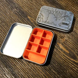 Puede incluir: Una caja metálica rectangular abierta con un interior naranja brillante dividido en seis compartimentos. La tapa de la caja presenta un diseño de mapa y el texto "CEIBA OUTFITTERS". La caja está sobre una superficie de madera.